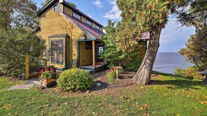 A yellow wooden house with a porch, a small garden, and a view of the water. - Lake Champlain Cottage w/ Private Deck & Fire Pit! (North Hero)