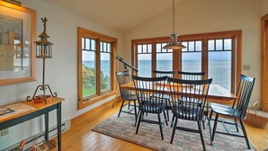 A dining area with a wooden table and chairs, large windows with a view of the sea, and a hanging light fixture. - Lake Champlain Cottage w/ Private Deck & Fire Pit! (North Hero)