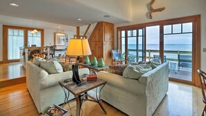 A spacious living area with a large window overlooking the sea, a wooden staircase, and a cozy seating arrangement. - Lake Champlain Cottage w/ Private Deck & Fire Pit! (North Hero)
