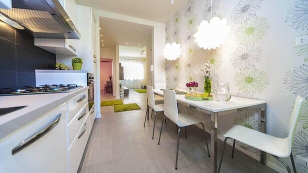 Une cuisine moderne avec des armoires blanches, une cuisinière à gaz et une hotte en acier inoxydable. On y trouve également un coin repas avec une table blanche, des chaises et un papier peint à motifs floraux.