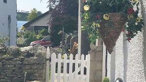 A white picket gate leading to a stone wall, a red car parked in the background, and a white building with a garden. - A beautiful family holiday cottage with lots of character in an idyllic village (Ulverston)