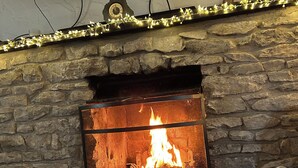 A stone fireplace with a lit fire, a television mounted above it, and a table with a candle and a book titled "PASS THE ROSE". - A beautiful family holiday cottage with lots of character in an idyllic village (Ulverston)