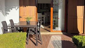 Un patio al aire libre con mesa y sillas de madera, una planta en maceta y una puerta corrediza de vidrio que da a un espacio interior con muebles blancos.
