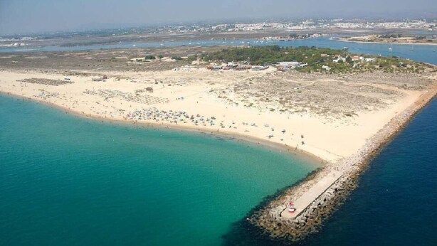 Luftaufnahme einer Küstenregion mit Sandstrand, einem steinernen Wellenbrecher und einer kleinen Insel mit Gebäuden.