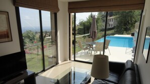 Ein modernes Wohnzimmer mit einem Glastisch, einer schwarzen Ledersofa und einem Fernseher. Große Fenster bieten Blick auf einen Swimmingpool und Gartenmöbel.