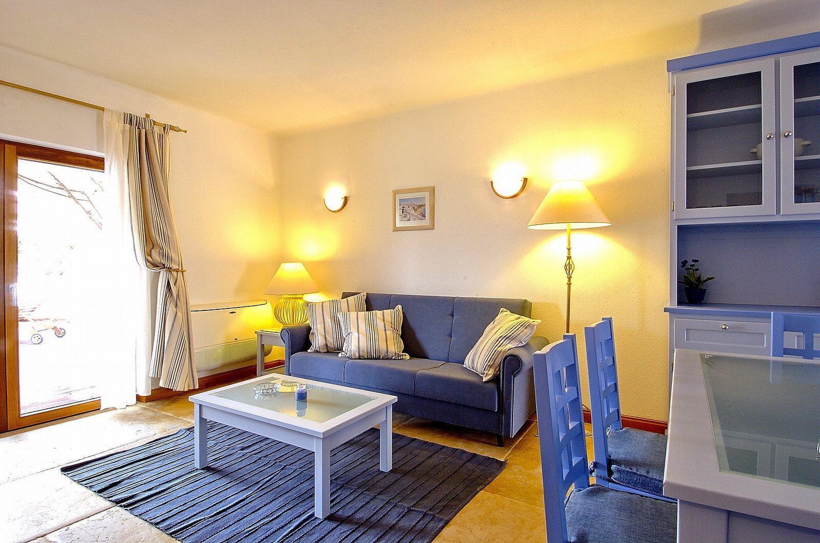 A living room with a sofa, a coffee table, and two lamps. There is a cabinet with glass doors and a small plant on top.