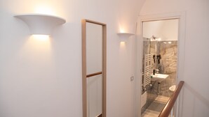 Un couloir avec une porte en bois et une porte en verre donnant sur une salle de bain dont le mur est recouvert de marbre et qui est équipée d’un lavabo blanc.