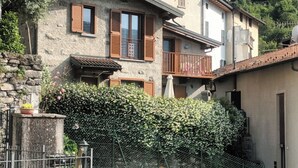 Un bâtiment traditionnel en pierre, doté de volets en bois, d’un balcon et d’une haie verdoyante et luxuriante.
