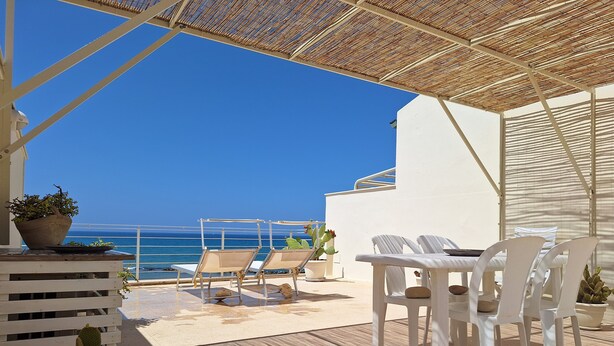 Una terraza con vista al mar, con una mesa y sillas, plantas en macetas y una barandilla blanca.