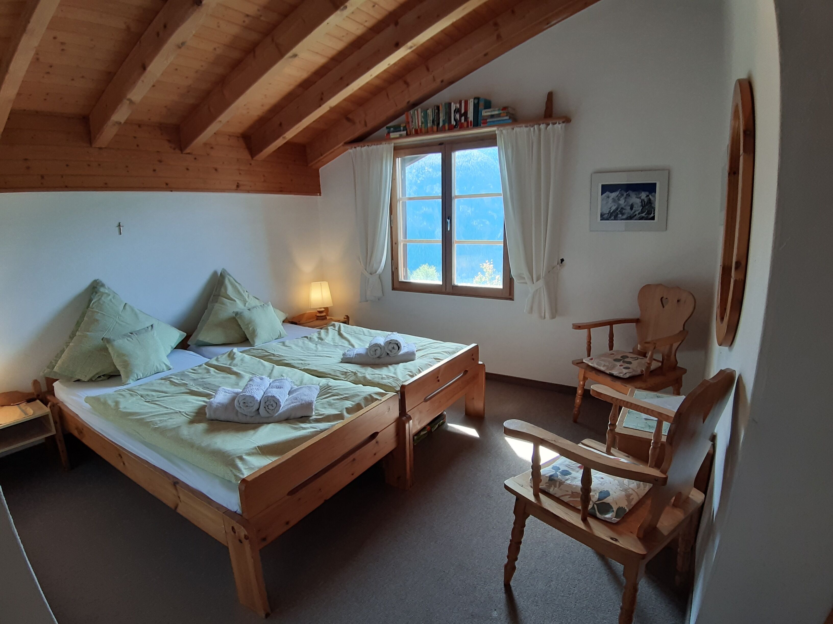 A cozy room with a wooden bed, a small wooden chair, a nightstand with a lamp, and a window with curtains.