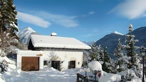 A snow-covered building with a wooden door and windows, surrounded by snow-laden trees and mountains in the background.