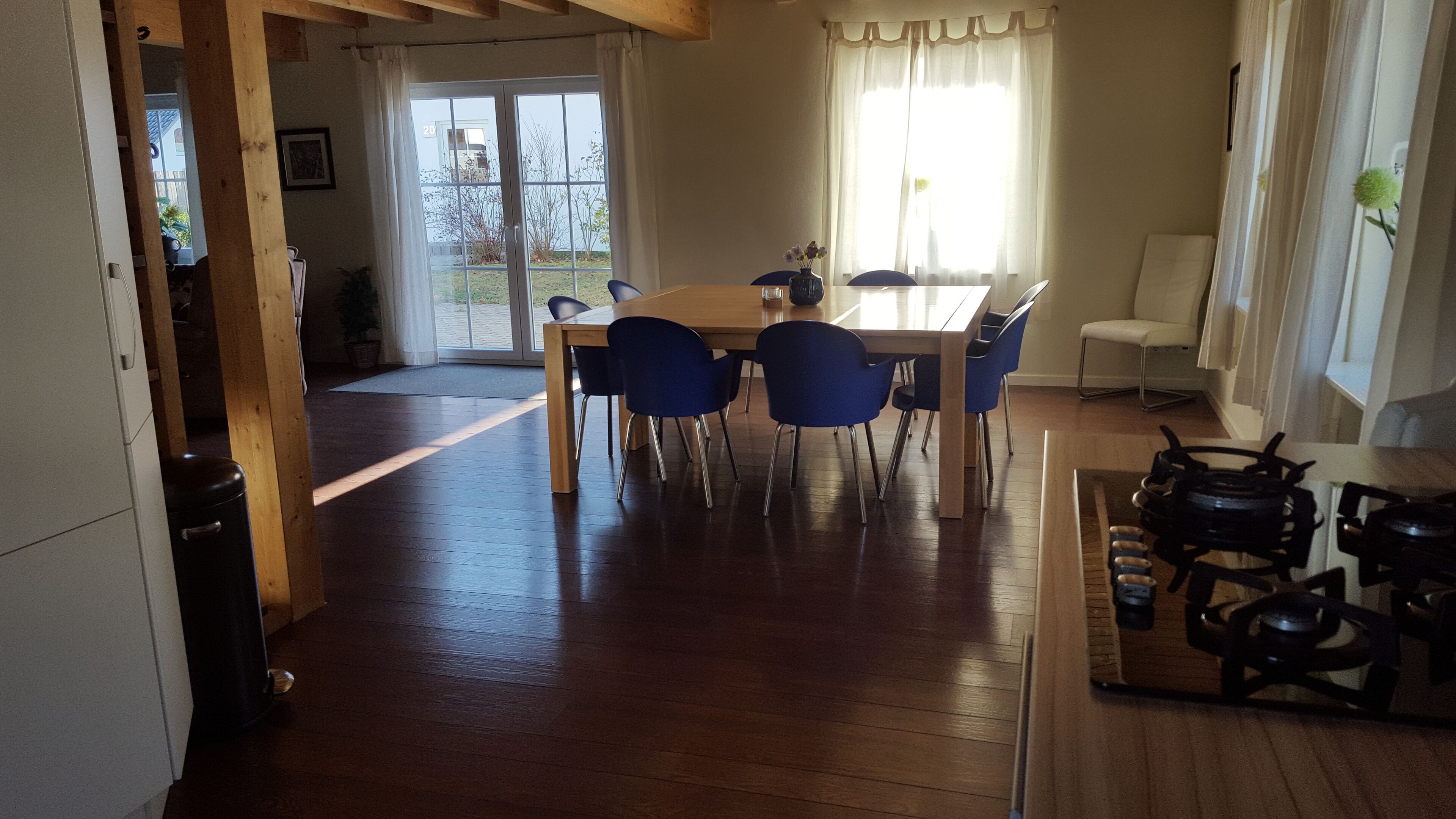 Un'area pranzo con tavolo in legno e sedie blu, una cucina con fornello e una porta scorrevole in vetro che dà su un balcone.