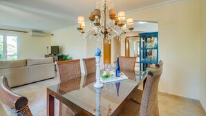 A dining area with a glass table, wicker chairs, a chandelier, and a blue cabinet with glassware.