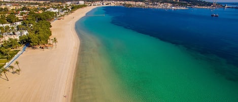 An aerial view of a coastal area with a sandy beach, clear blue waters, and a mountainous backdrop.