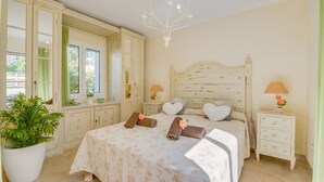 A bedroom with a large bed, two bedside tables, a chandelier, a potted plant, and a window with curtains.
