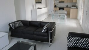 Een moderne woonkamer met een zwarte bank, een glazen salontafel en een eethoek met transparante stoelen en een witte tafel.