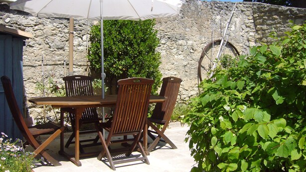 En udendørs terrasse med et bord, stole, en hvid parasol og en stenvæg i baggrunden.