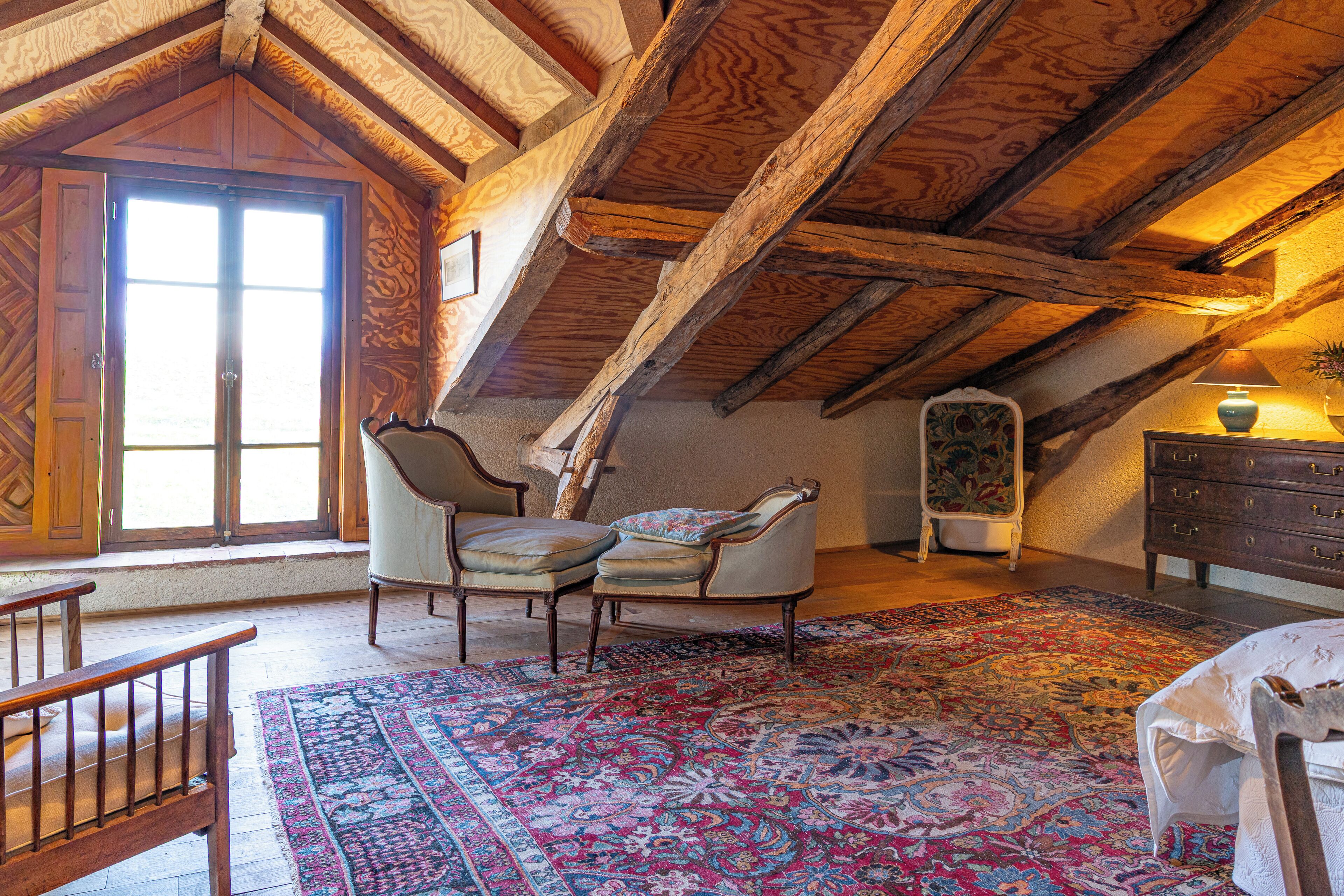 Une pièce mansardée spacieuse avec des poutres apparentes, une grande fenêtre, un tapis à motifs et un fauteuil classique.