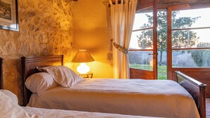 Un dormitorio con pared de piedra, cama de madera, lámpara de noche y ventana con cortinas.