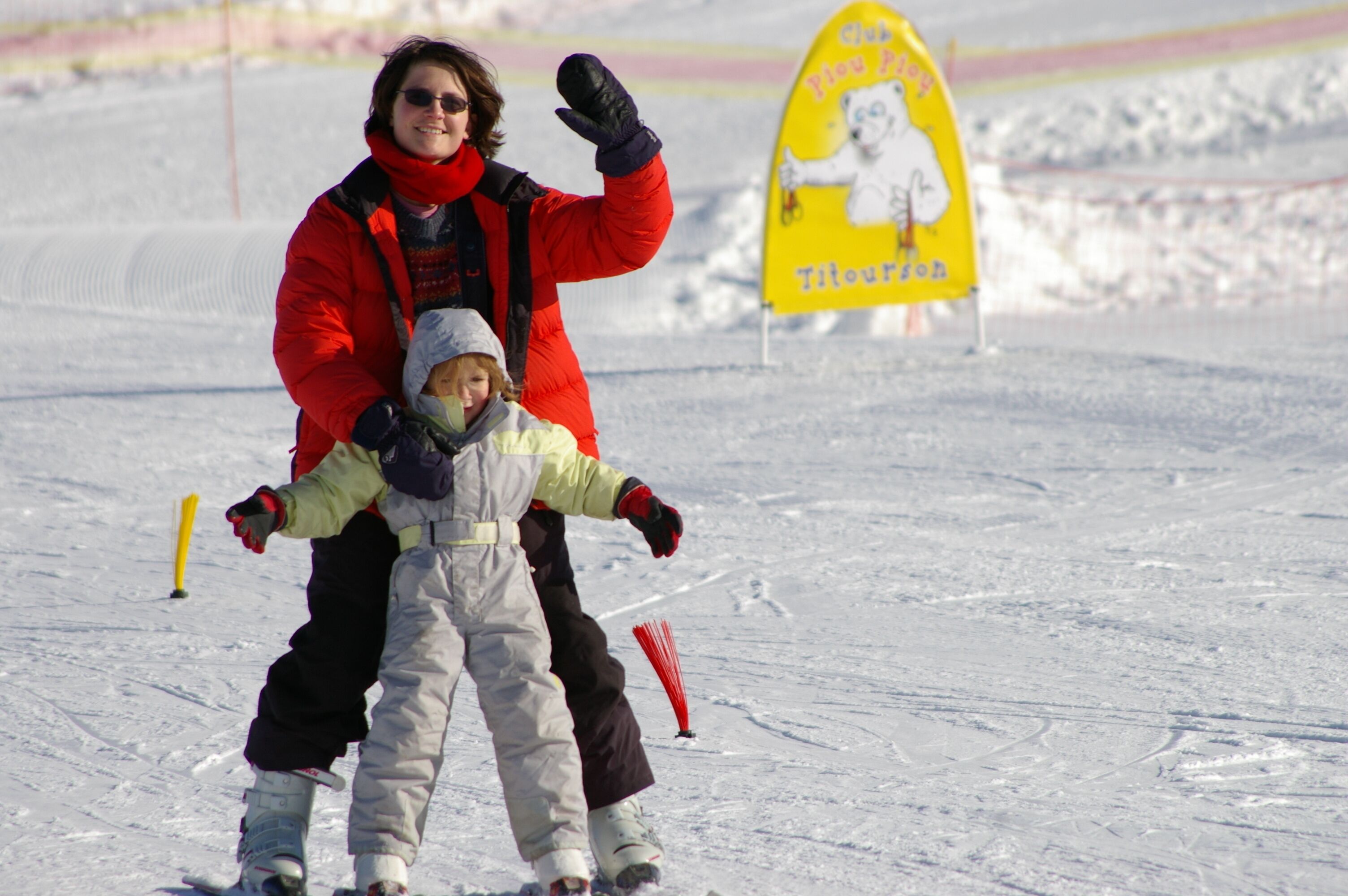 A person in a red jacket and black pants standing on a snowy surface with a yellow sign in the background.