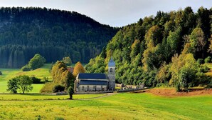 A rural church with a bell tower, surrounded by lush green fields and dense forested hills.