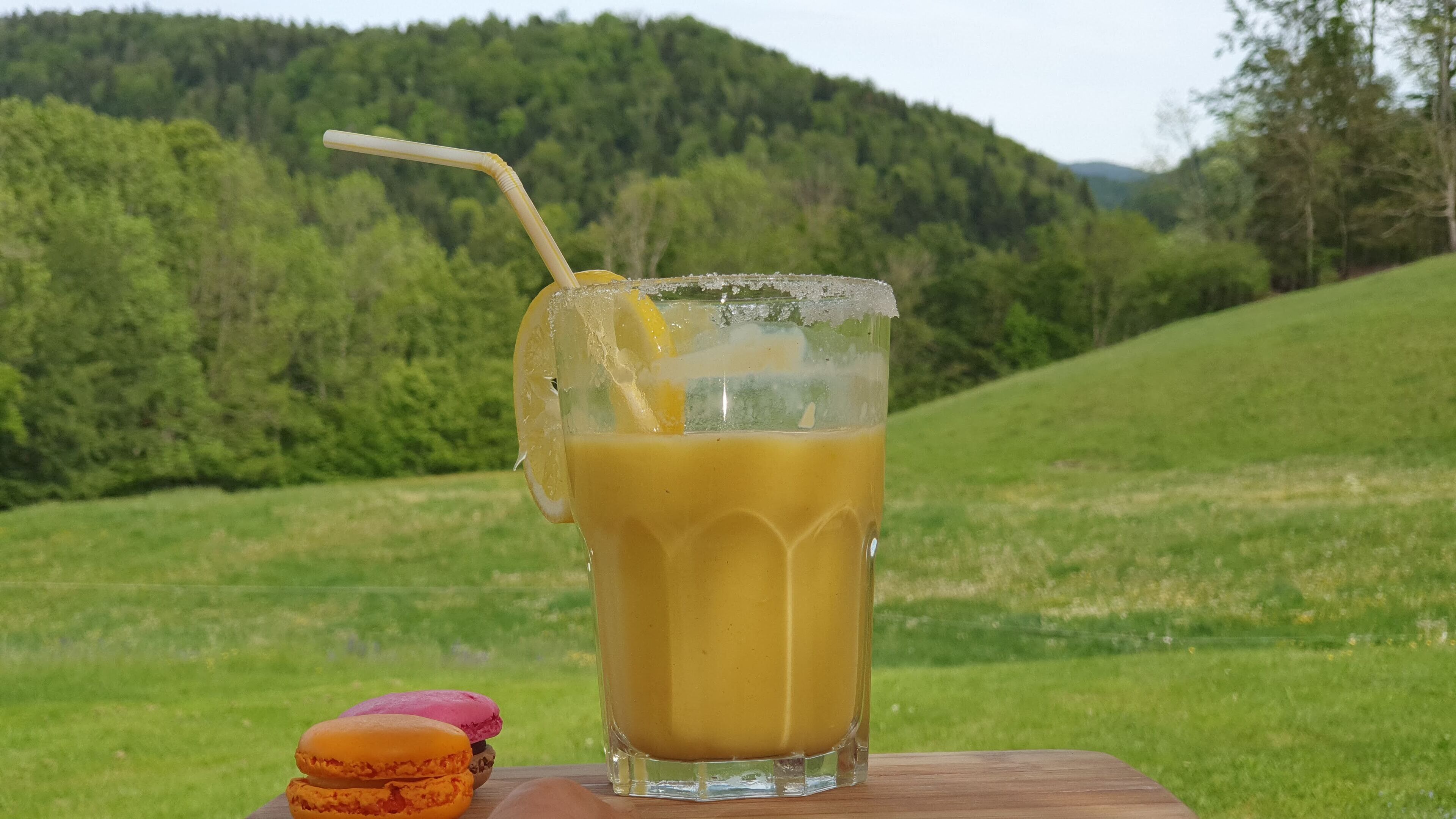 A glass of iced coffee with a straw and lemon slice on a wooden table. In the background, there is a grassy field and a forested hill.