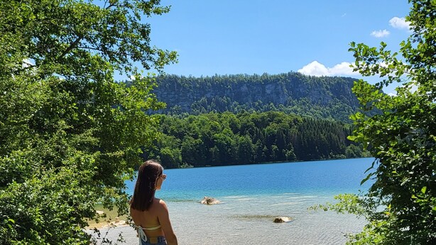 A person standing on a grassy area overlooking a clear blue lake with a forested hill in the background.
