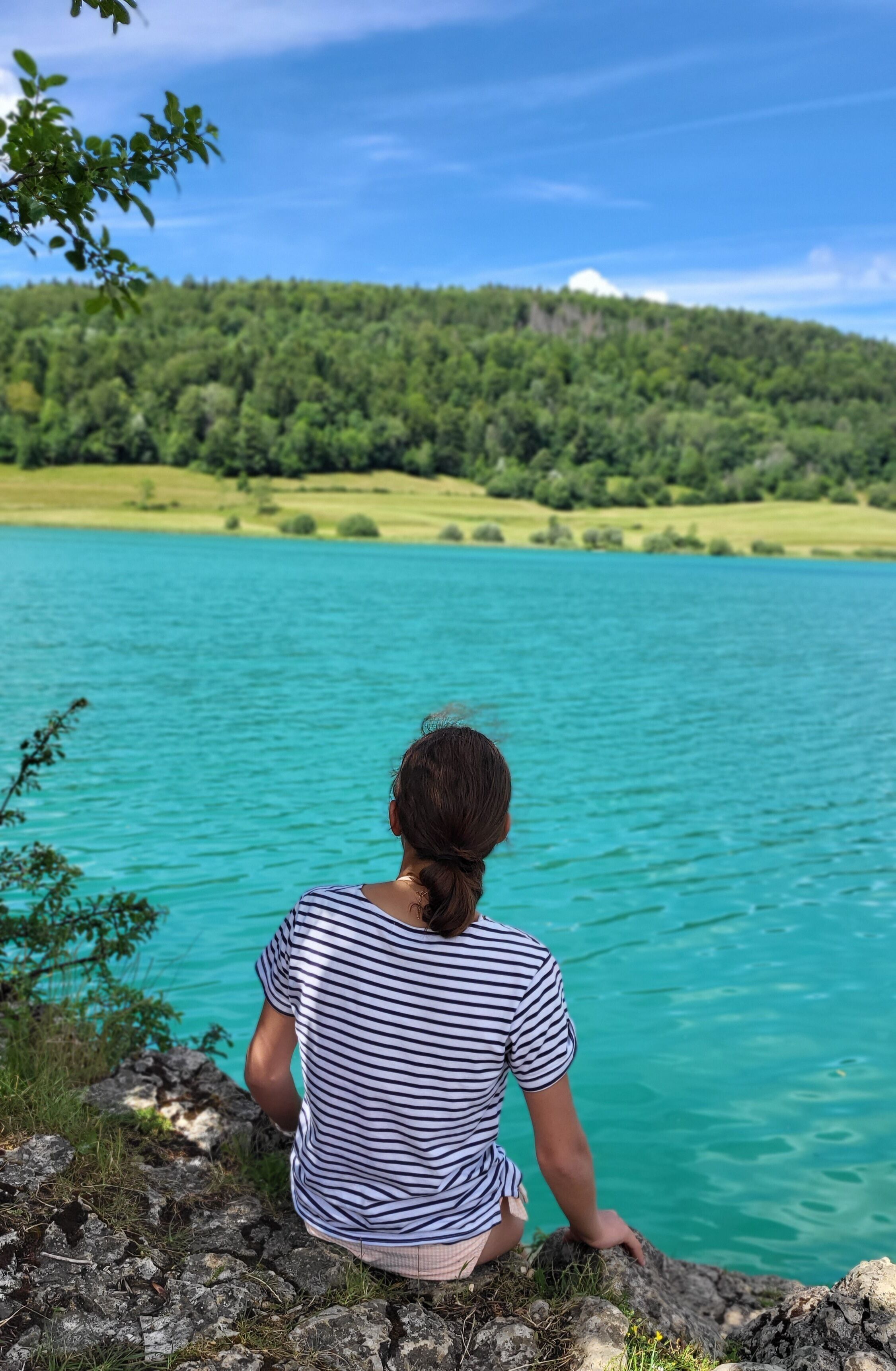 A person sitting on a rocky ledge overlooking a turquoise lake with a forested hill in the background.