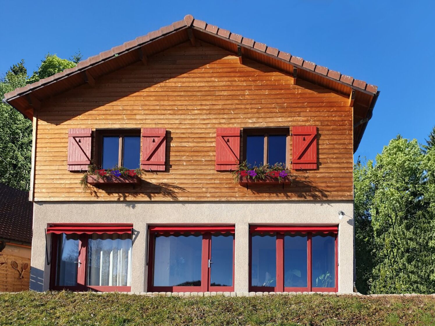 A two-story wooden house with red shutters, large windows, and a sloped roof.