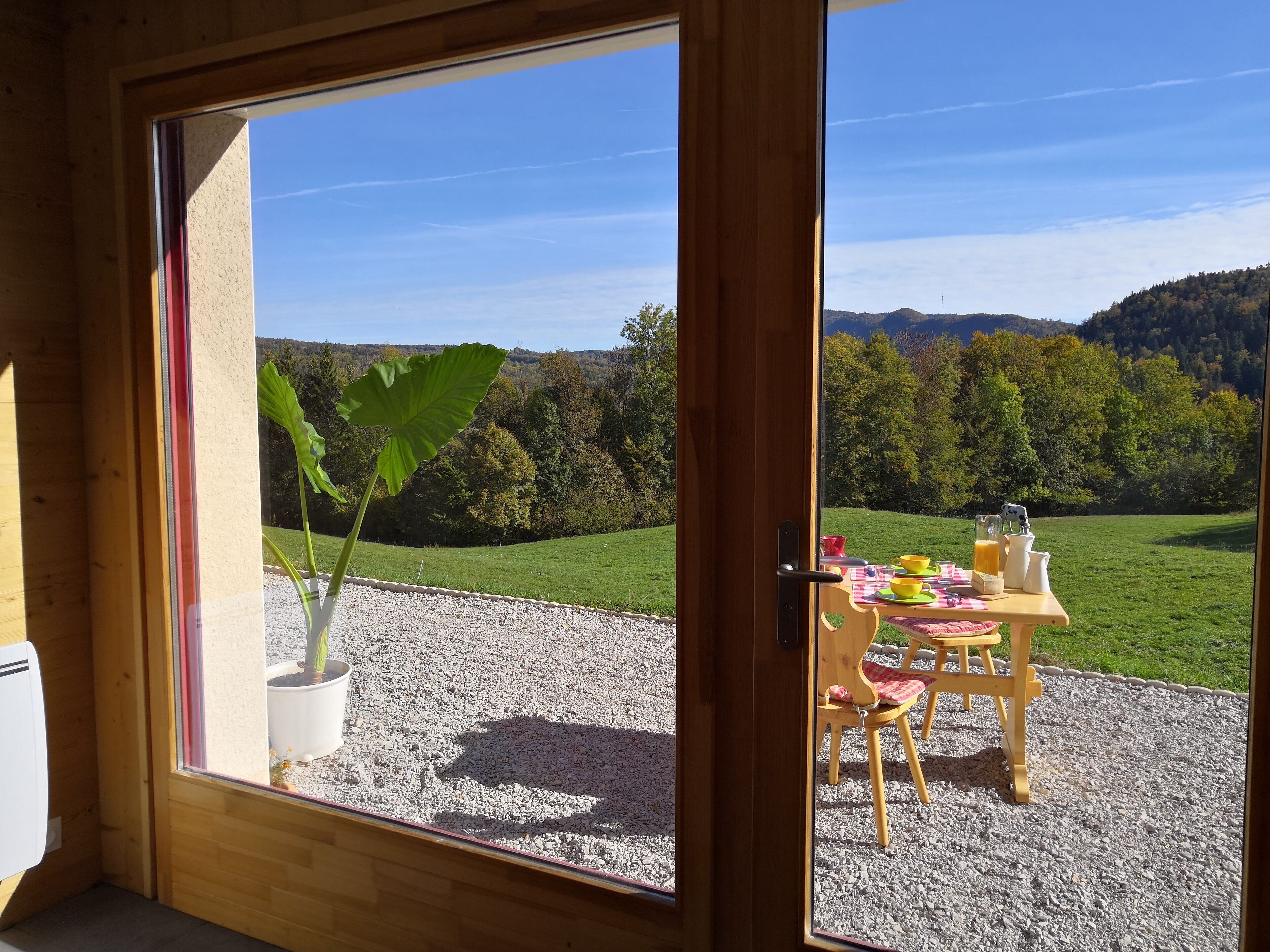 Une pièce avec une porte en bois donnant sur un patio où une table est dressée pour le petit-déjeuner, agrémentée d’une plante en pot, et d’où l’on peut admirer un paysage verdoyant avec des collines.