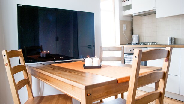A dining area with a wooden table and chairs, a flat-screen TV, and a kitchen in the background. - Sopot Sleeps - Sopot Suite (Sopot)