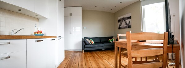 A compact kitchen with white cabinets, a wooden countertop, and a bowl of fruit. A dark sofa and a wooden dining table with chairs are in the living area.