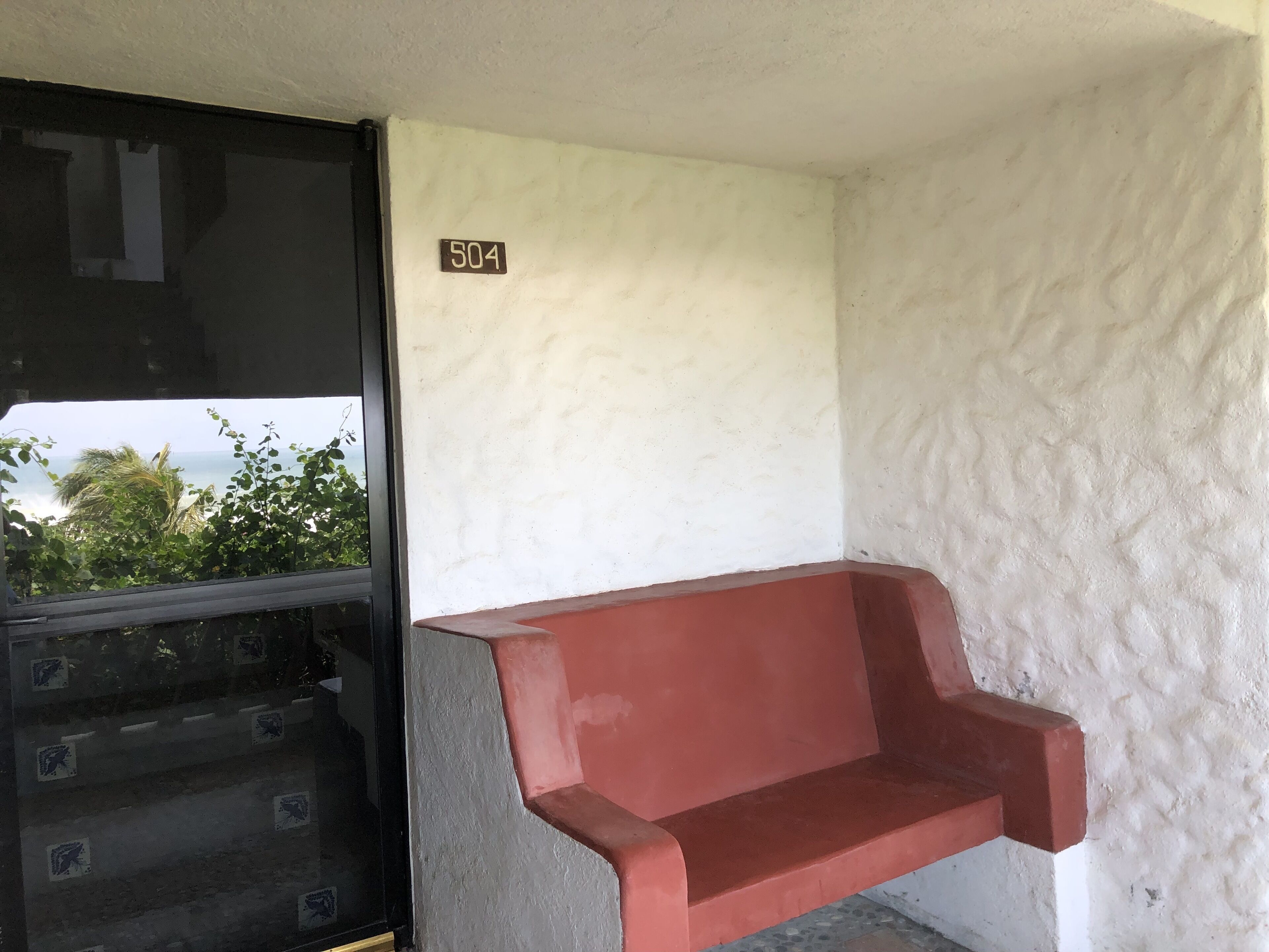 Een kamer met een rode bank, een witte muur met een bordje met het nummer '504', en een raam met uitzicht op groen en de zee.