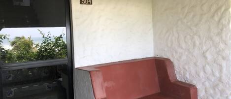 Een kamer met een rode bank, een witte muur met een bordje met het nummer '504', en een raam met uitzicht op groen en de zee.