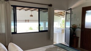 Une chambre à coucher avec un lit, une fenêtre avec des rideaux, une table de chevet avec un vase, et une vue sur l’extérieur.