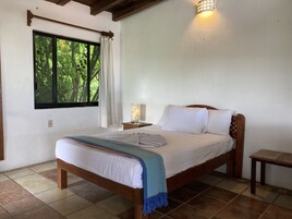 Un dormitorio con una cama de madera, ropa de cama blanca, un plaid azul, una mesita de noche con una lámpara y una ventana con cortinas.
