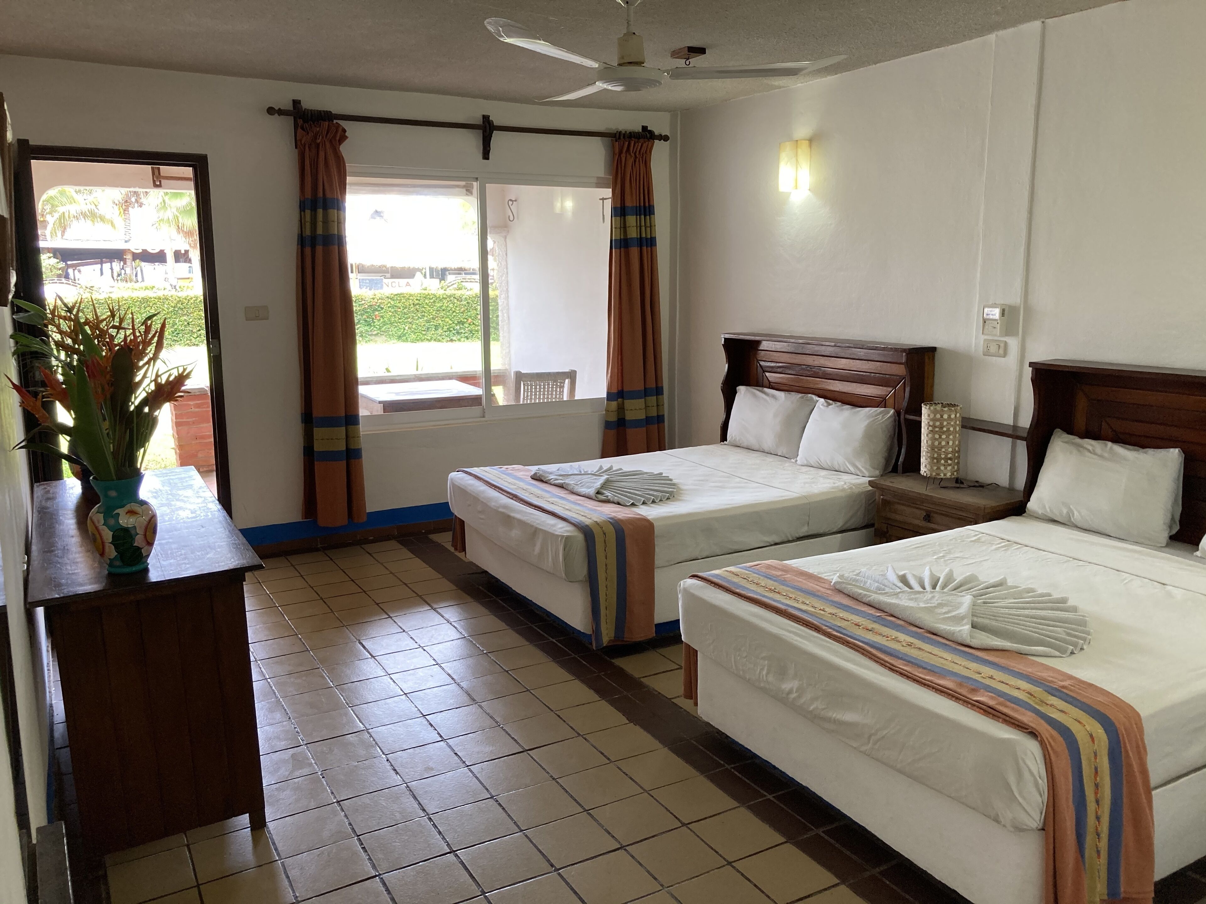 A hotel room with two beds, a wooden headboard, a bedside table, a nightstand, a window with curtains, a ceiling fan, and a view of the outside.