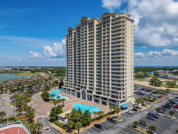 A high-rise hotel with multiple balconies, a swimming pool, and a parking lot.