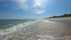 A beach with waves gently lapping onto the shore, a clear sky, and a distant view of a hilly landscape. - OCEAN VIEWS! Immaculate Quintessential Nantucket Beach Cottage see virtual tour. (Nantucket)