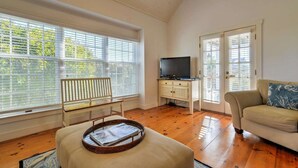 A spacious living room with wooden floors, a white sofa, a television on a cabinet, and large windows with blinds. - OCEAN VIEWS! Immaculate Quintessential Nantucket Beach Cottage see virtual tour. (Nantucket)