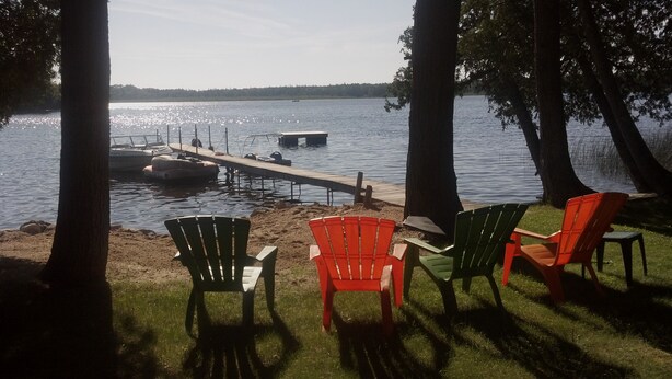 A lakeside view with a dock extending into the water, surrounded by trees. There are several Adirondack chairs on the grassy area.