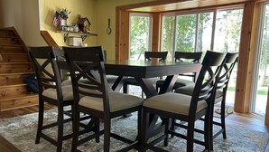 A dining area with a wooden table and chairs, a rug, a staircase, and a window with a view of trees.