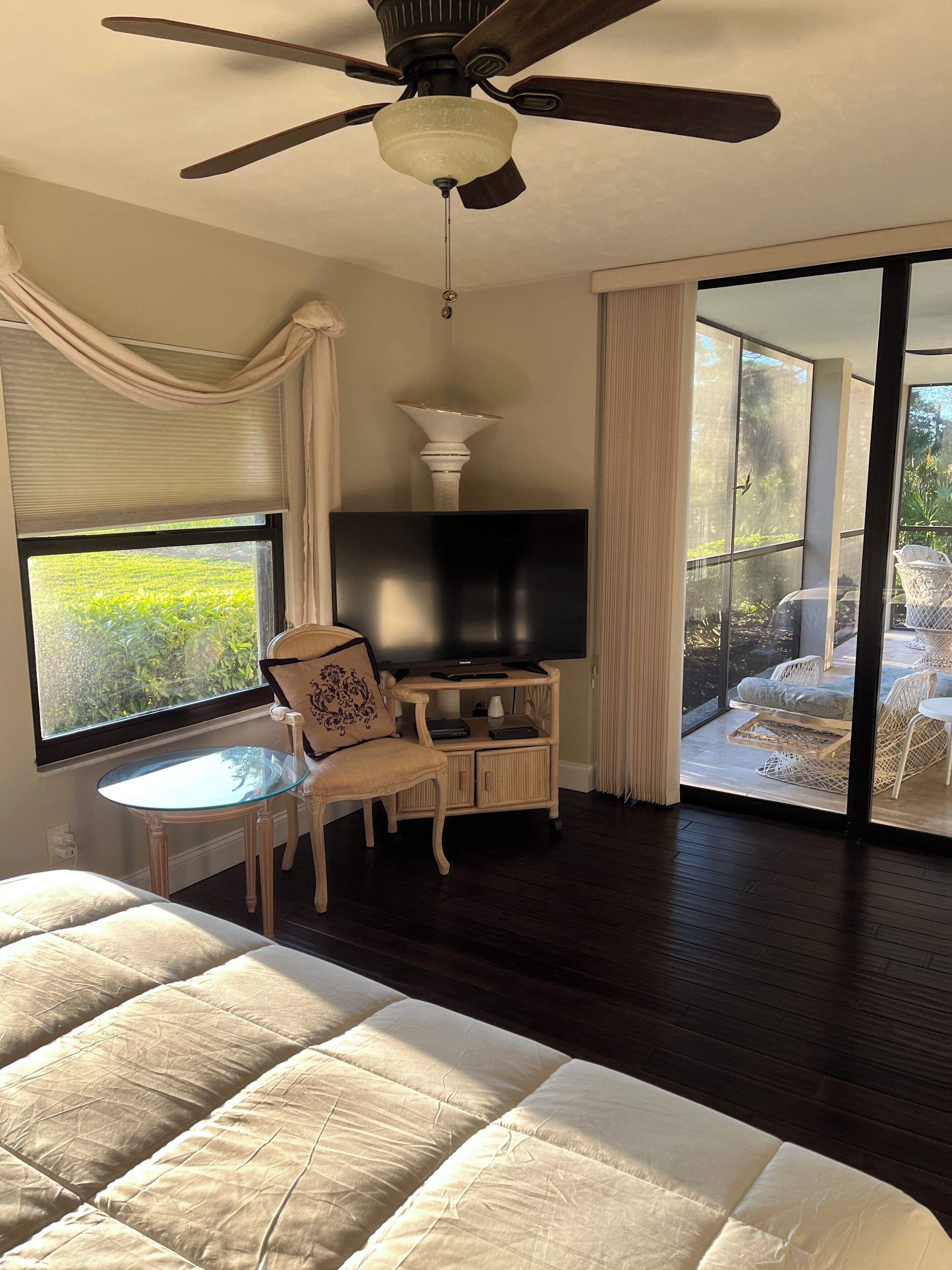 A bedroom with a large bed, a TV on a wooden stand, a small table, and a chair. There is a balcony with wicker furniture and a view of greenery.