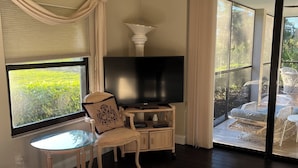 Un dormitorio con una cama grande, un televisor sobre un soporte de madera, una mesita pequeña y una silla. Cuenta con un balcón amueblado con mobiliario de mimbre y vistas a la vegetación.