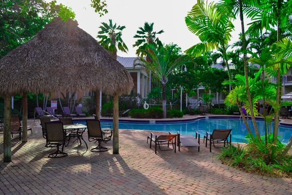Un espace aménagé au bord de la piscine, avec des chaises longues, un gazebo à toit de chaume et une piscine entourée de palmiers.