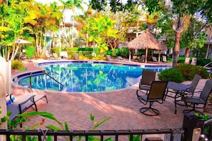 A swimming pool with lounge chairs and a thatched roof structure, surrounded by lush greenery and a brick patio. - Luxury Condo-FREE PARKING,WIFI,BIKES,BIG POOL,WALK TO RESTAURANTS, BEACH  CHAIRS (Key West)