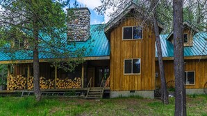 A rustic cabin with a stone chimney, wooden exterior, and a porch with stacked firewood. - Sun Mountain Ranch Bunkhouse - Near Crater Lake (Fort Klamath)