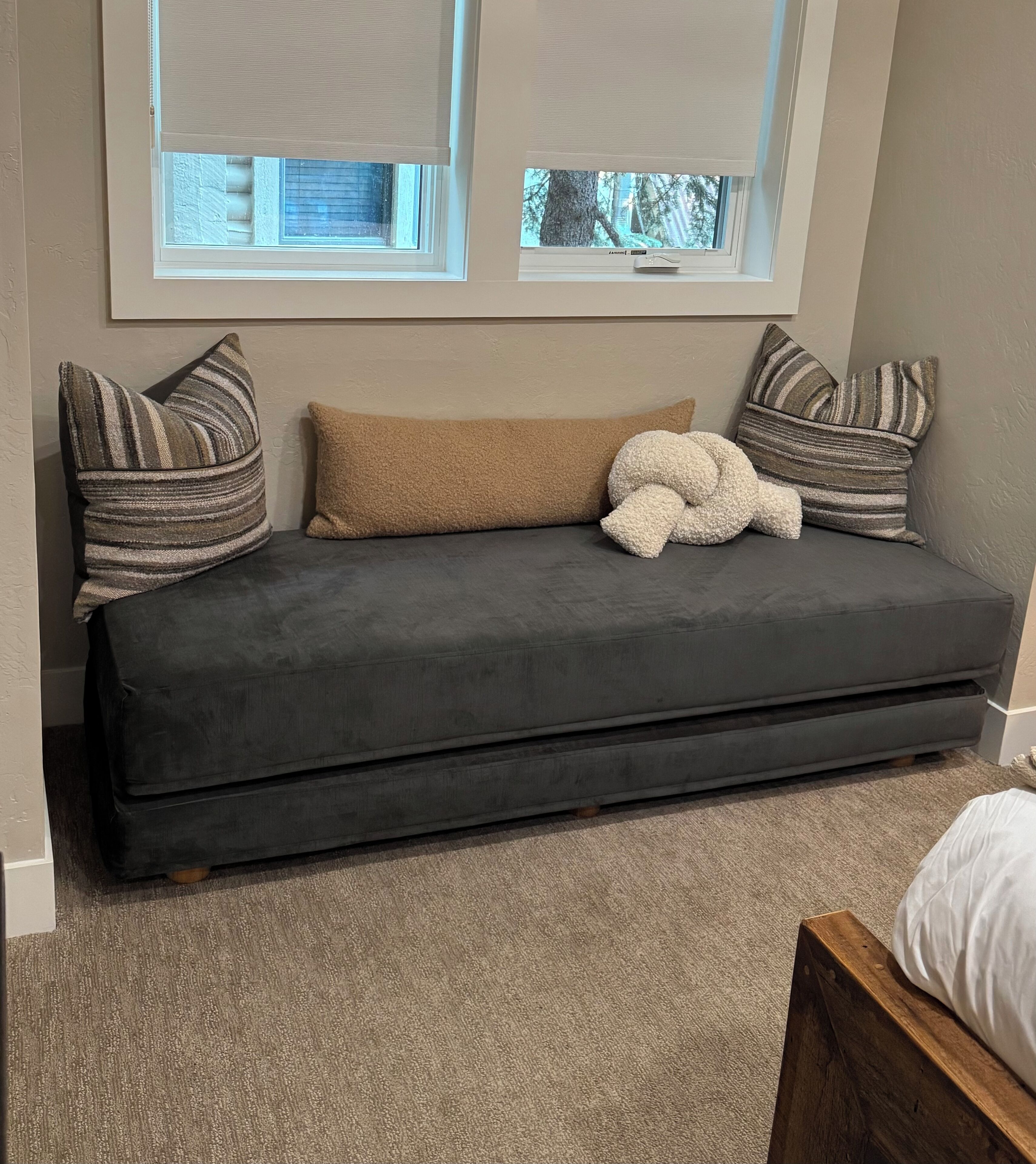 Un canapé gris avec un coussin de couleur beige et deux coussins à rayures, le tout placé contre un mur percé de deux fenêtres.