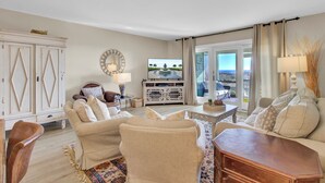 A spacious living room with a large TV, comfortable seating, and a view to the outside. - Award-Winning Resort Amenities | Mariners Walk 1B (Isle of Palms)