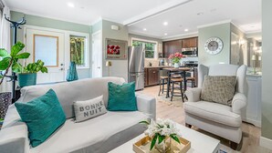 Uma sala de estar moderna com um sofá, poltronas e uma mesa de centro. Uma cozinha é visível ao fundo, com uma área de refeições e um relógio na parede.